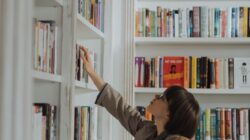 woman in beige coat standing in front of white wooden book shelf